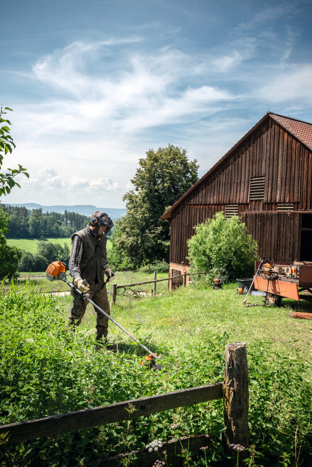Man using STIHL FS230 brushcutter for sale to trim grass near a wooden barn in rural area