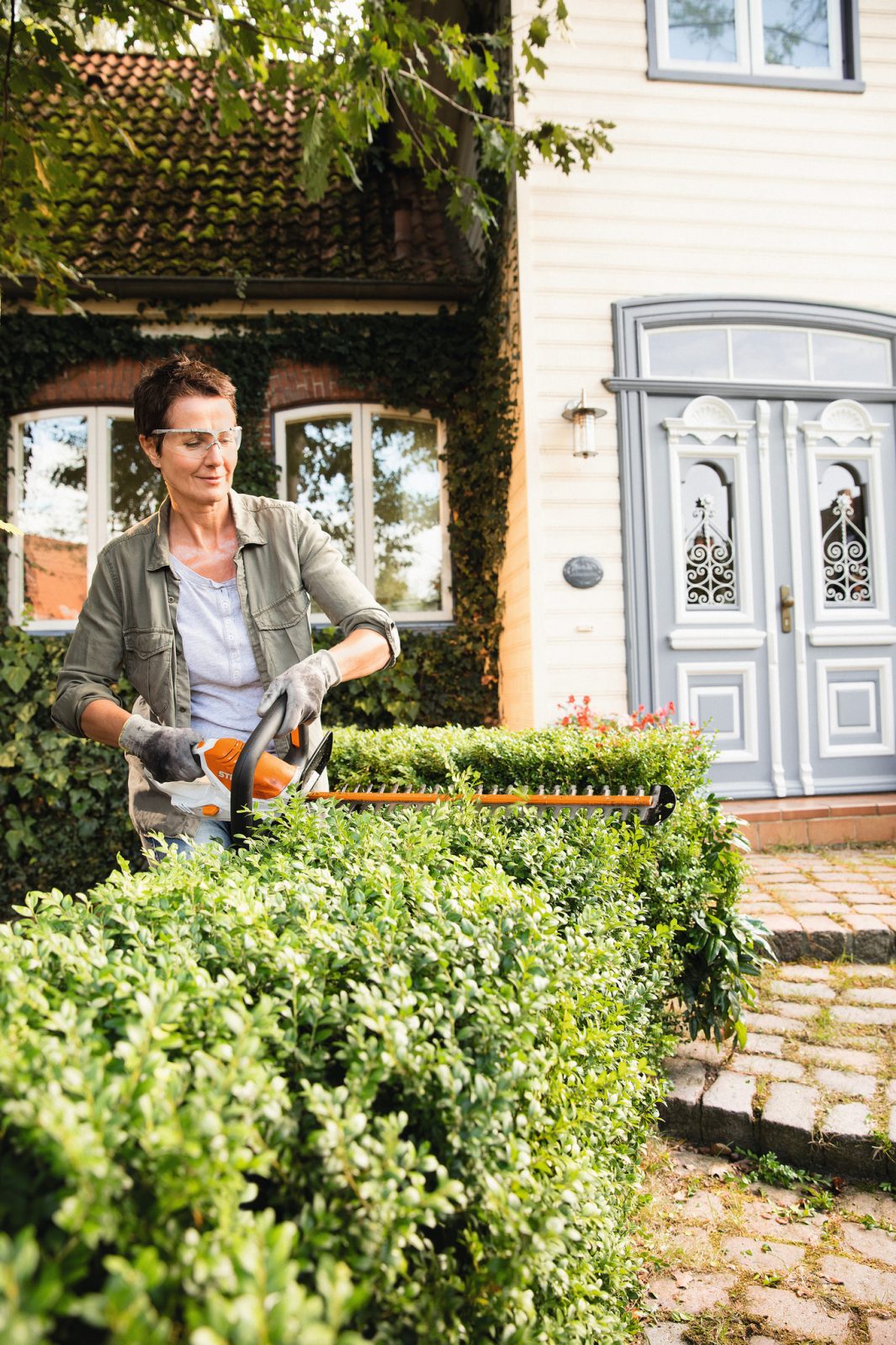 Person trimming garden hedge with STIHL HSA45 hedge trimmer outside a classic house