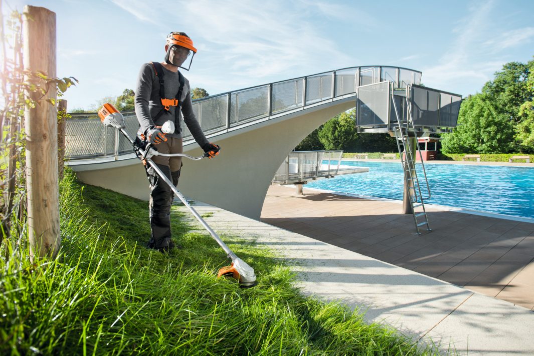 Person using STIHL FSA90 brushcutter for sale to trim grass near a pool and bridge on a sunny day