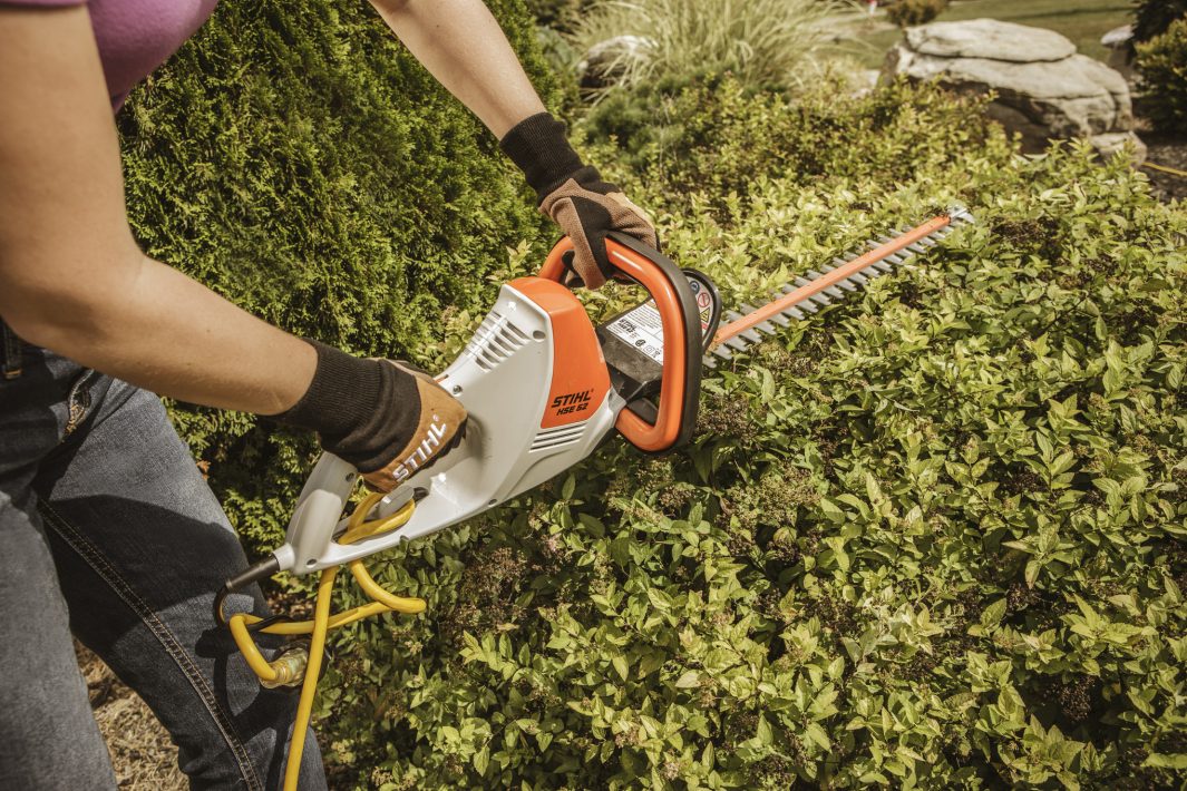Person using STIHL HSE52 hedge trimmer to trim green bushes in garden