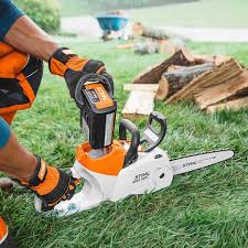 STIHL MSA160 C-B chainsaw in white and orange being charged on grass with wood logs in background